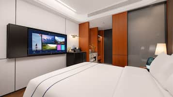 A modern hotel room with a large bed, a flat-screen TV mounted on the wall, a desk with a lamp, and a wardrobe.