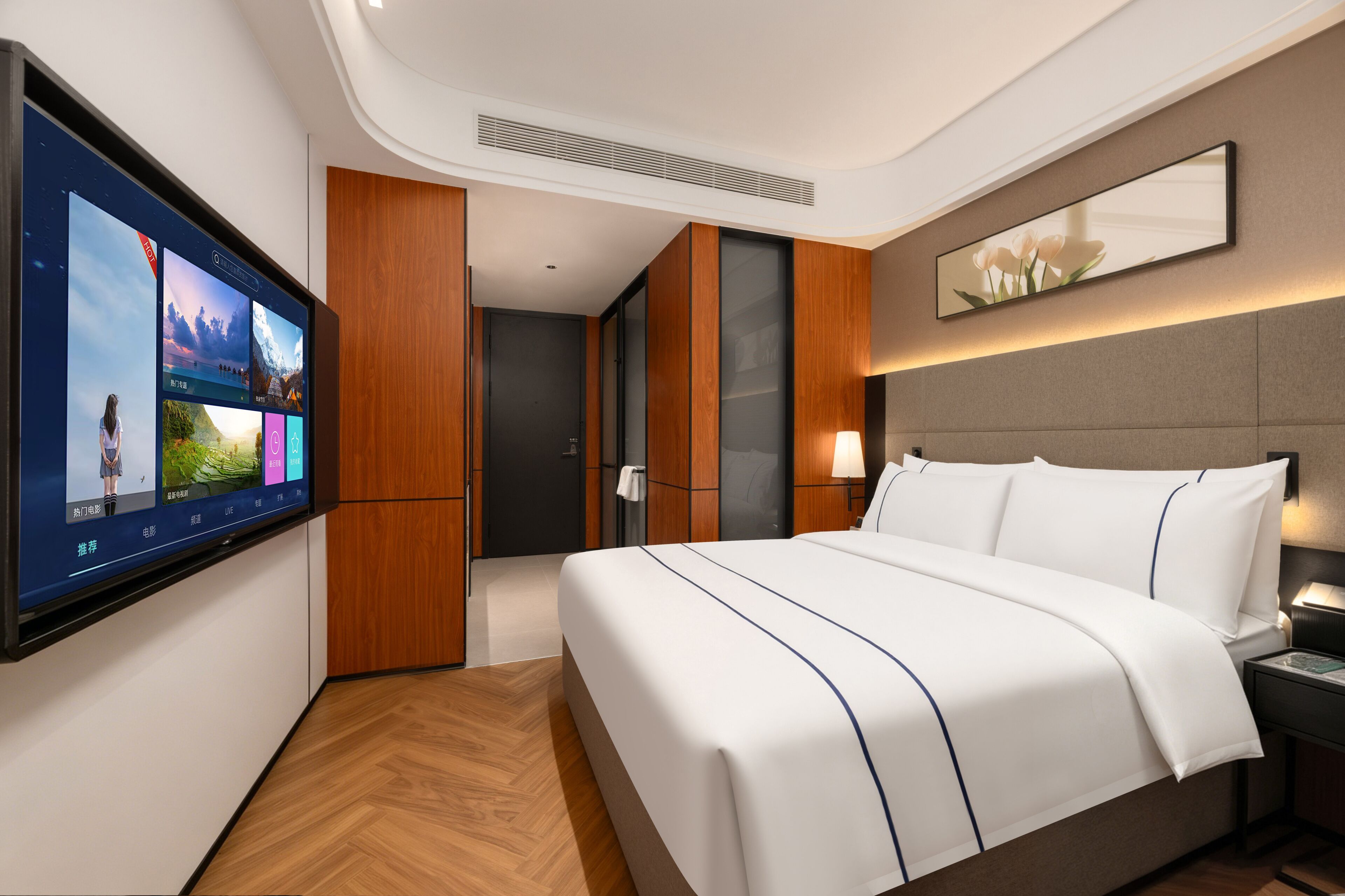 A hotel room with a large flat-screen TV mounted on the wall, a bed with white bedding, a nightstand, a lamp, and a mirror above the bed.