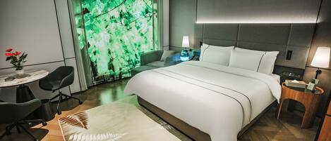 A modern hotel room with a large bed, a desk with a chair, a small table, and a wall-mounted lamp.