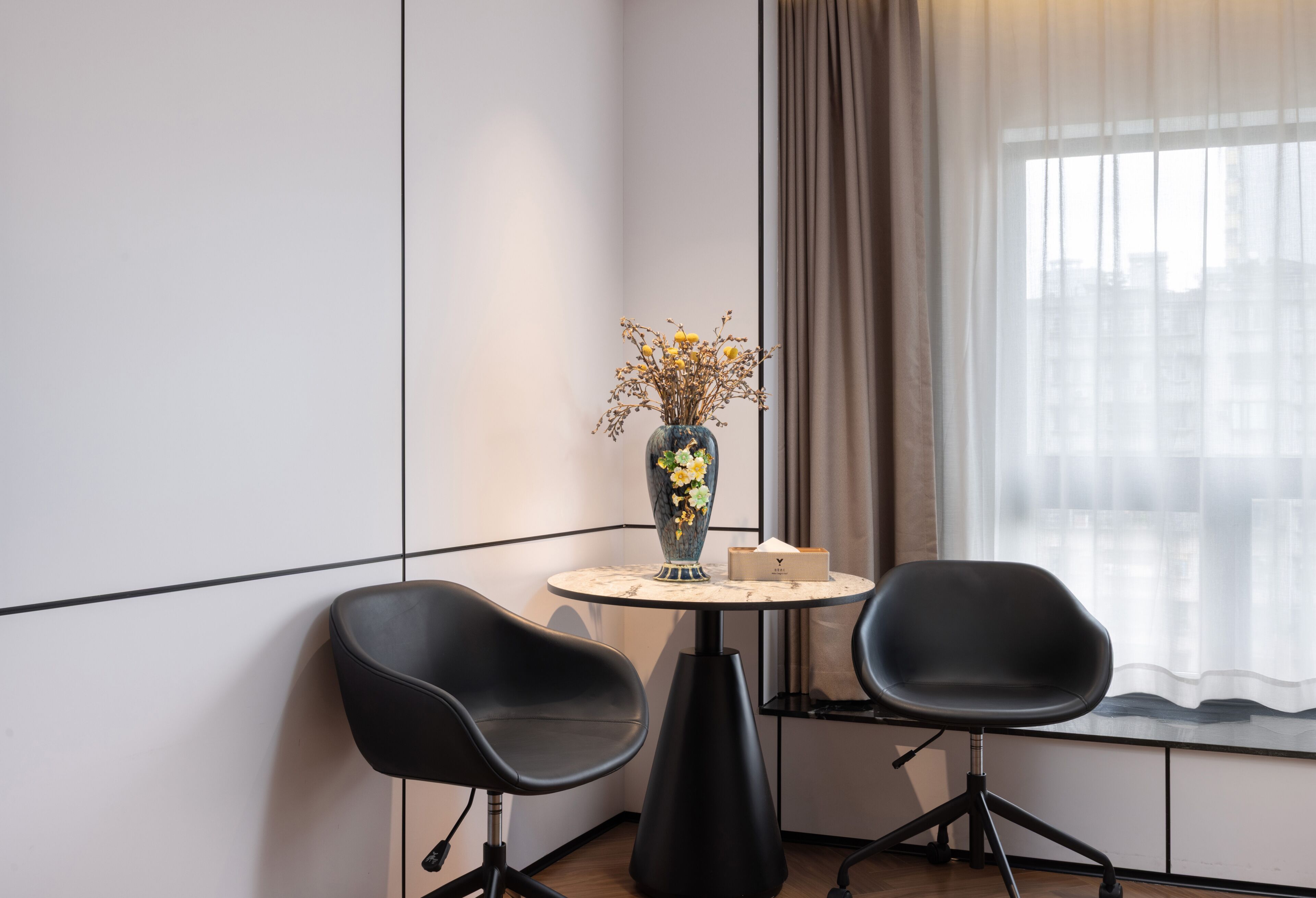 A modern room with a round table, two black chairs, a vase with flowers, and a view through a window with curtains.