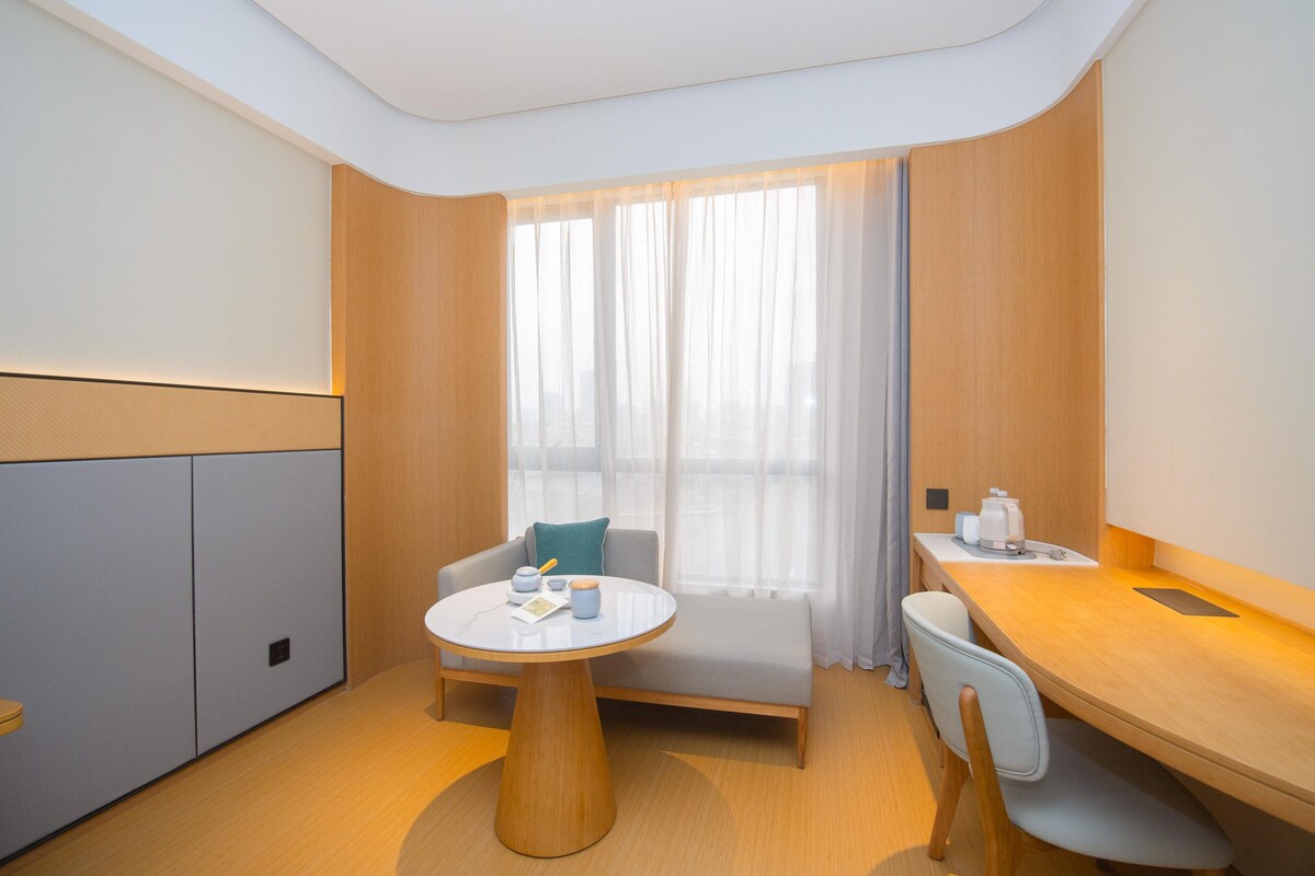 Une chambre d’hôtel moderne avec une table ronde en bois, un canapé, un bureau et une grande fenêtre ornée de rideaux légers.