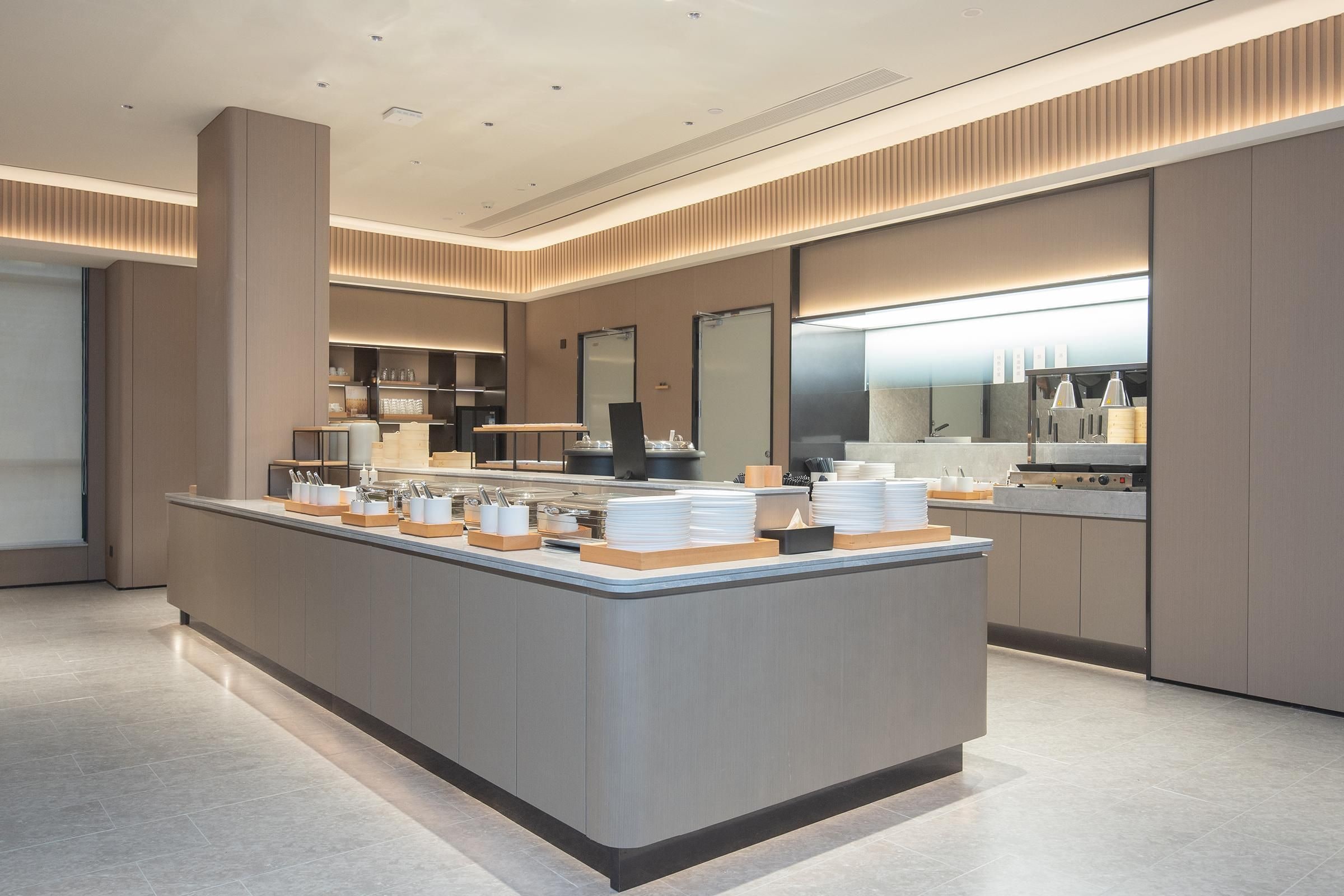 A modern hotel lobby with a central reception desk, a large mirror, and a kitchen area in the background.