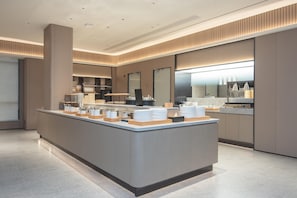 A modern hotel lobby with a central reception desk, a large mirror, and a kitchen area in the background.
