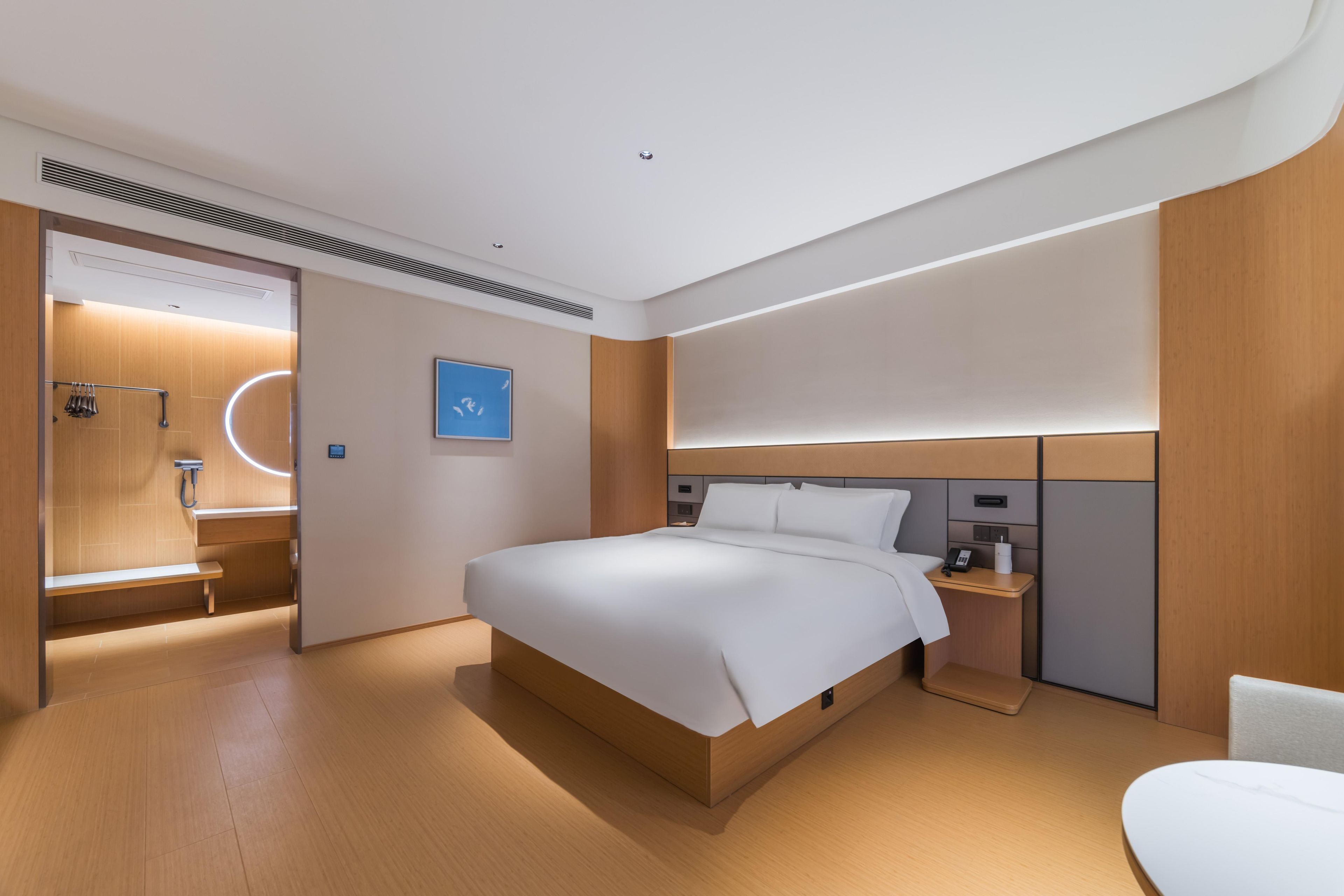 A modern hotel room with a wooden floor, a large bed with white linens, a bedside table with a lamp, and a small seating area.