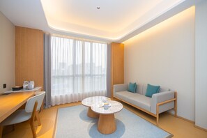A modern hotel room with a wooden desk, a round coffee table, a sofa with cushions, and a large window with sheer curtains.
