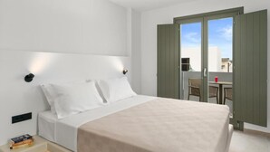 Quarto de hotel com uma cama, mesa de cabeceira com livros, janela com cortinas e varanda com cadeiras e uma mesa.
