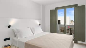 Quarto de hotel com uma cama, mesa de cabeceira com livros, janela com cortinas e varanda com cadeiras e uma mesa.