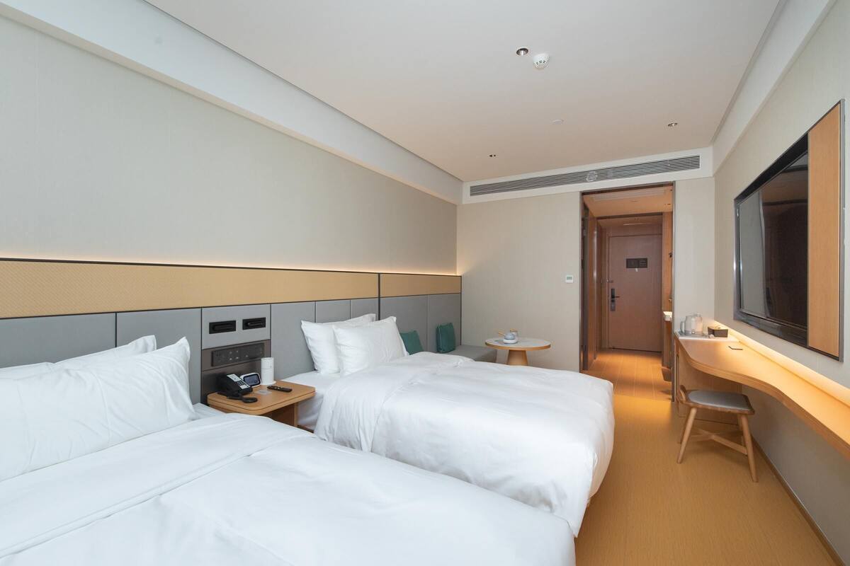 A hotel room with two beds, a desk, a chair, a small table, a mirror, and a door leading to another room.