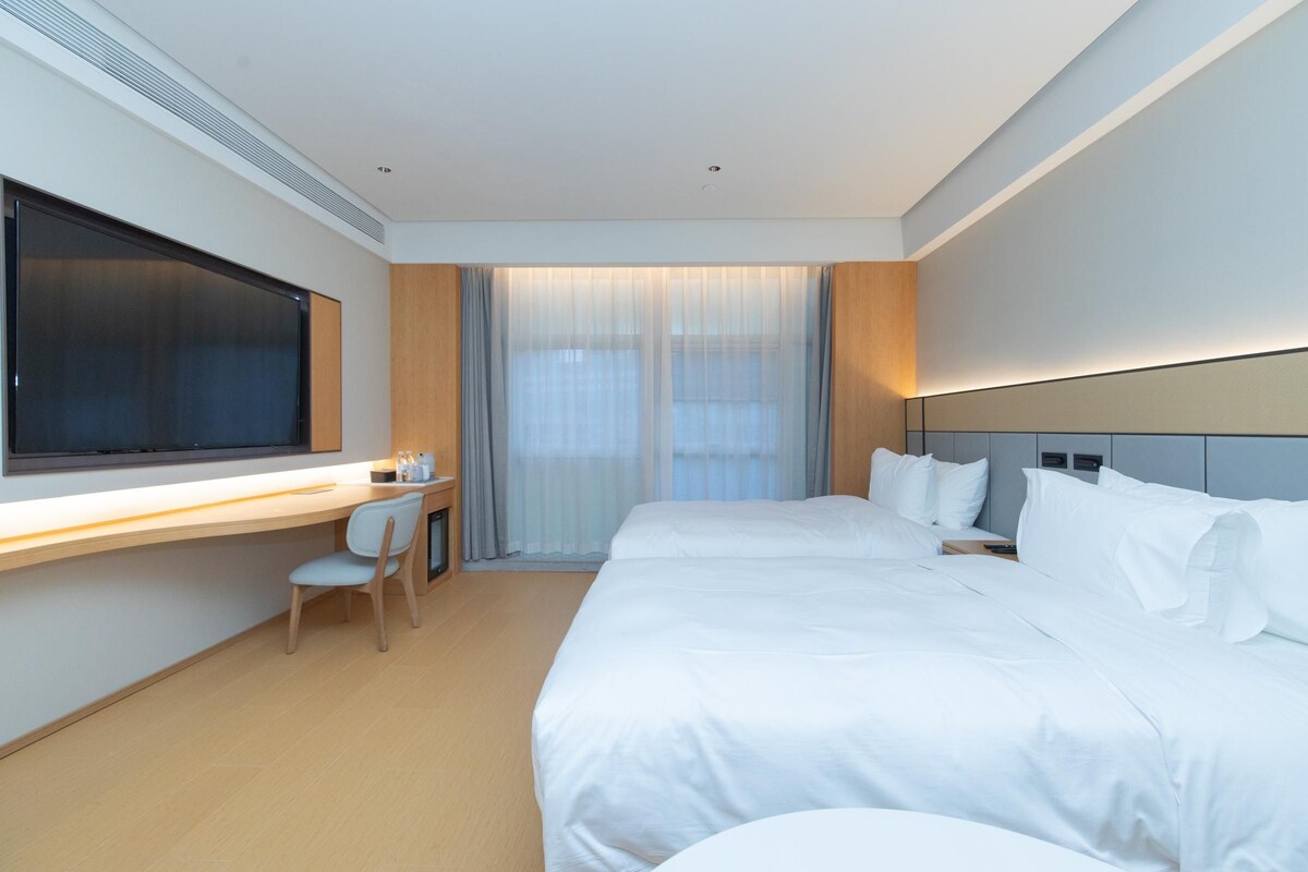 A modern hotel room with a large bed, a desk with a chair, and a large window with curtains.