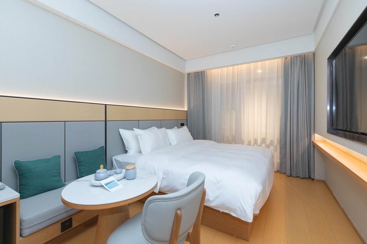 A modern hotel room with a neatly made bed, a small round table with a chair, and a TV mounted on the wall.