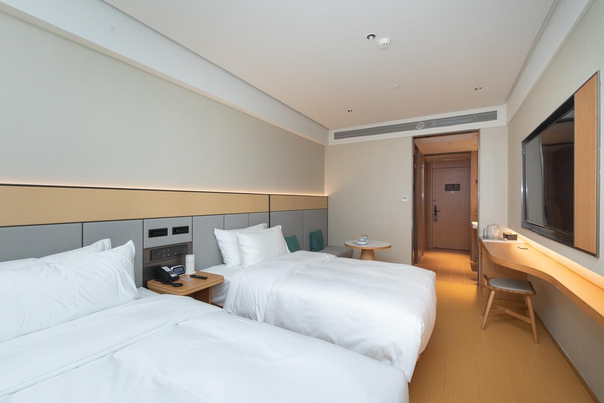 A hotel room with two beds, a desk, a chair, a small table, a mirror, and a door leading to another room.
