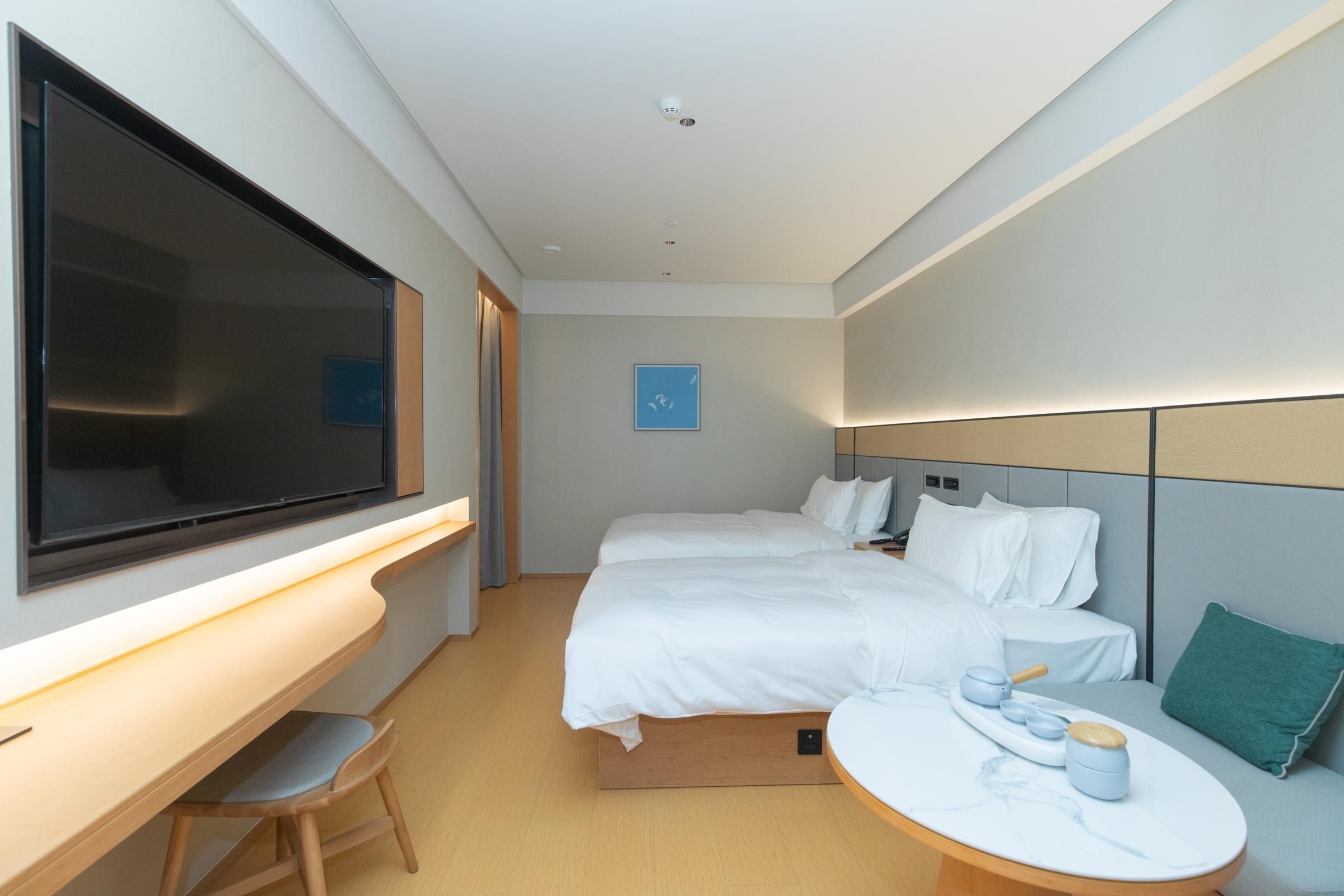 A modern hotel room with a large flat-screen TV, a desk with a chair, two beds with white bedding, a round table with a tray of items, and a green pillow on the right side.