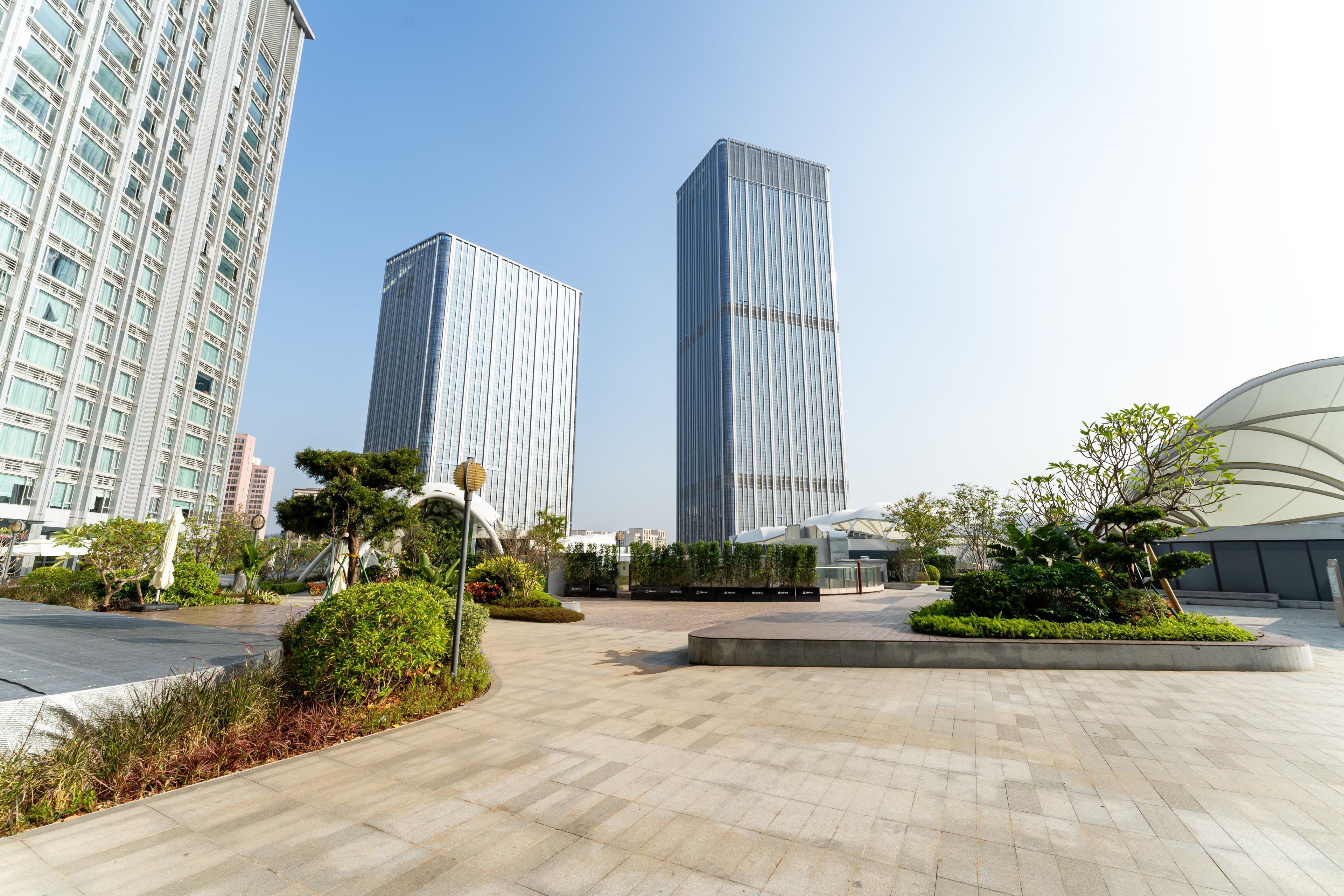 A modern urban plaza with two tall buildings, landscaped areas, and a curved structure.