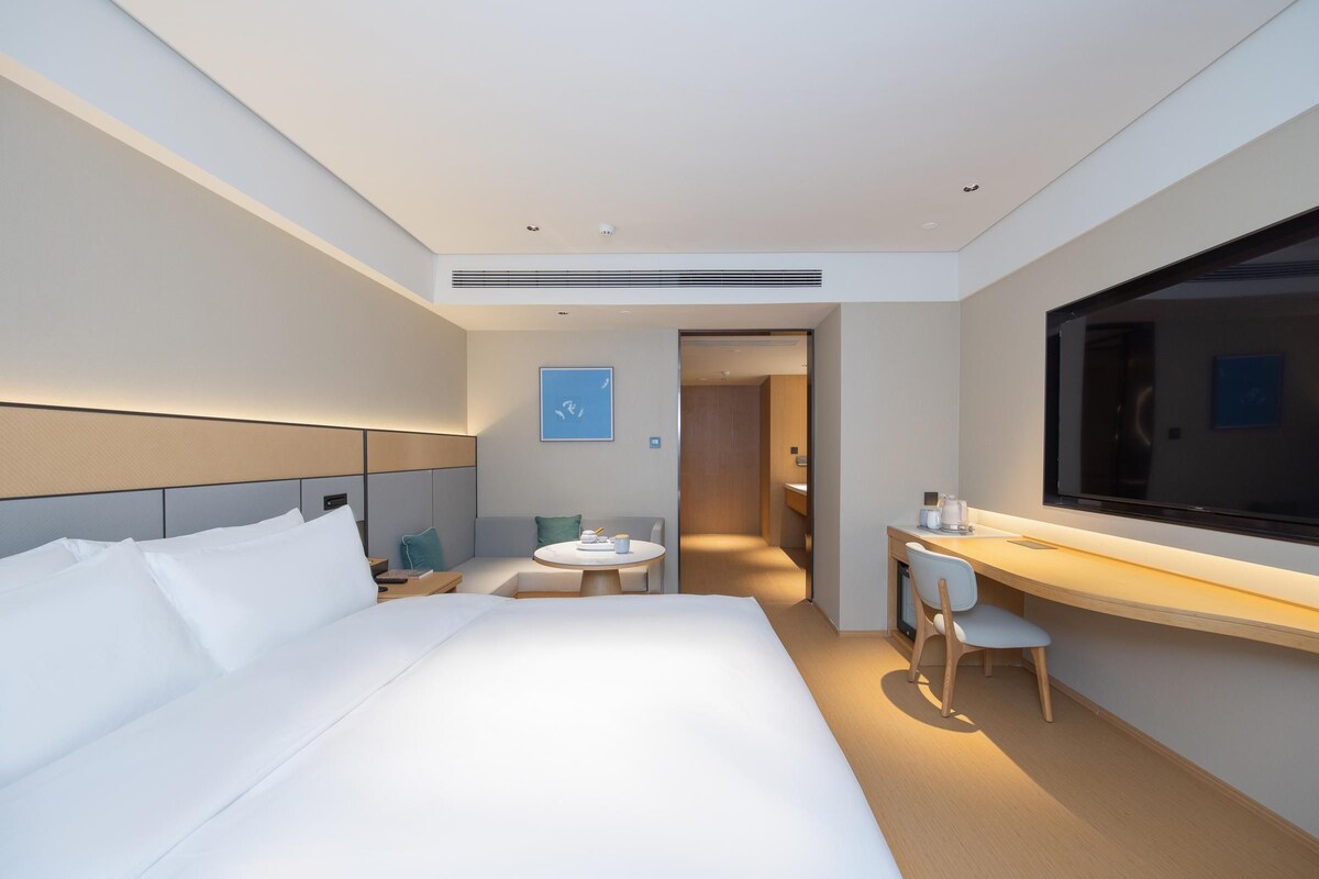 A modern hotel room with a large bed, a desk with a chair, a small table, and a TV.