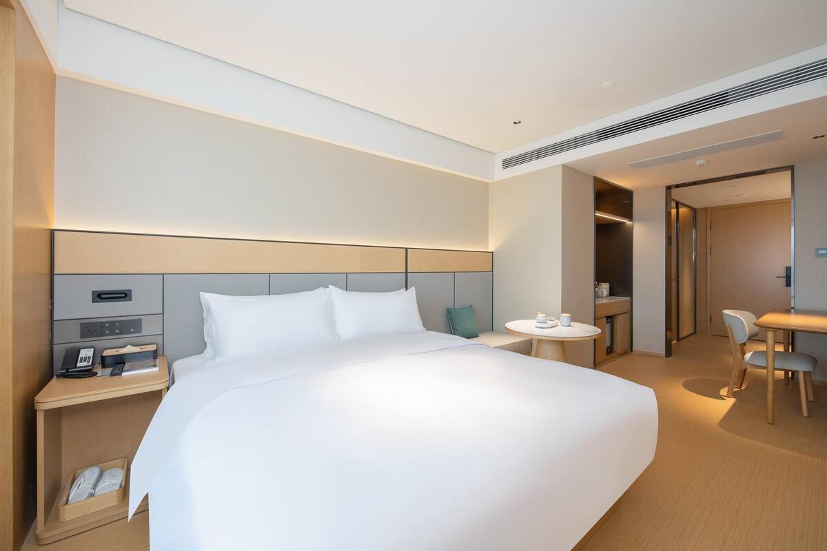 Una camera d'albergo moderna con un letto grande, un comodino con telefono e sveglia, un piccolo tavolino con una sedia, e un'area pranzo separata con tavolo e sedie.