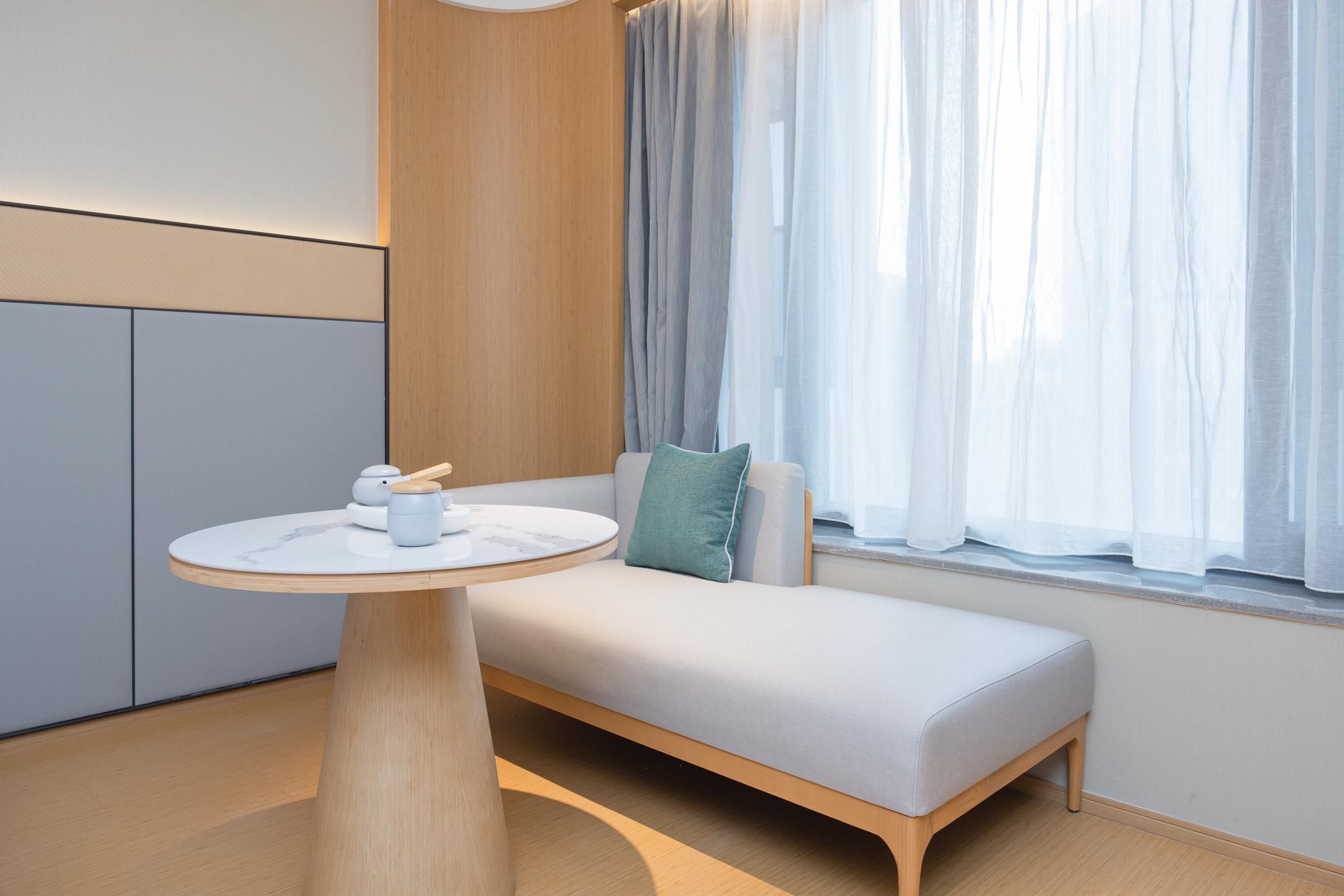 A modern room with a minimalist design, featuring a wooden bench, a round table with a white surface, and a window with sheer curtains.