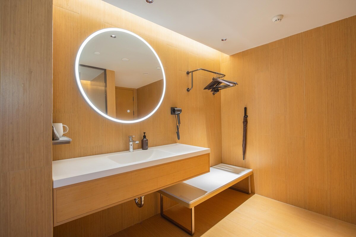 A modern bathroom with a wooden vanity, a round mirror with a light, a towel rack, and a handheld showerhead.
