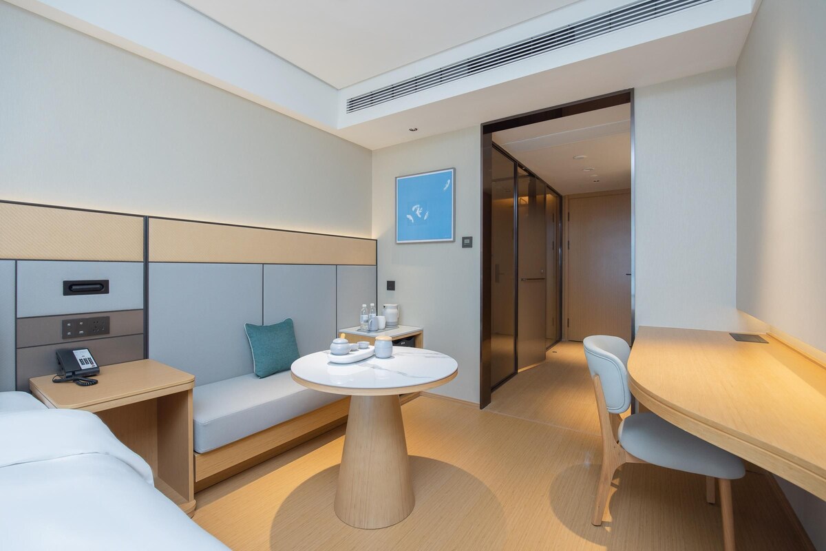 Una habitación de hotel moderna con una cama, una mesita de noche con teléfono, una mesa redonda con una silla, un sofá con un cojín y una obra de arte en la pared.