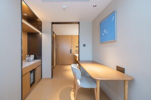 A modern, compact room with a wooden desk, a chair, a small kitchenette, and a door leading to another room.