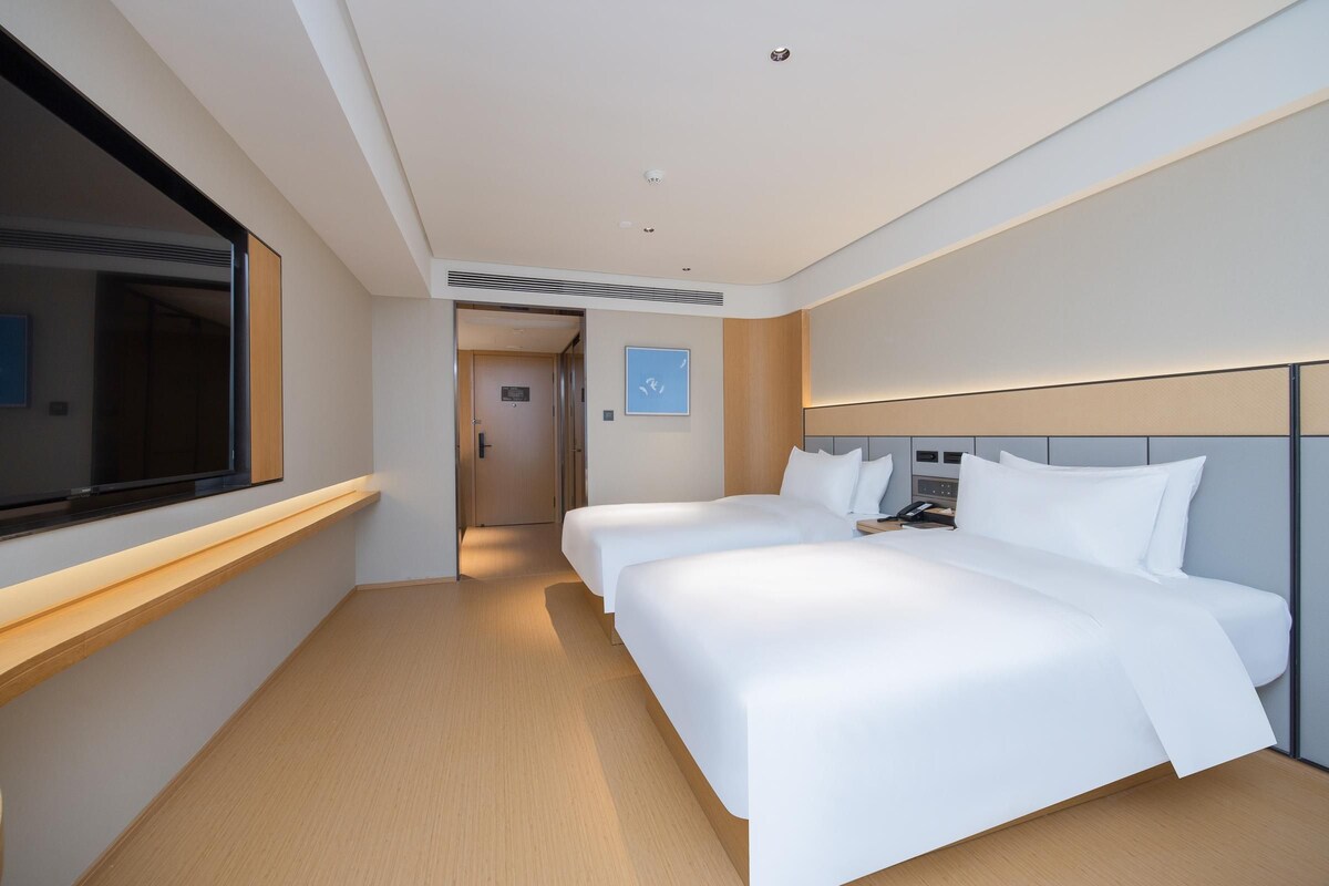 Habitación de hotel con dos camas, una mesita de noche con teléfono, una ventana y una puerta que da a otra habitación.