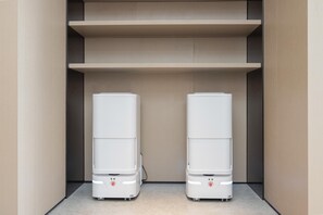 Una habitación moderna y minimalista con dos electrodomésticos blancos y compactos sobre el suelo, un estante de madera encima y una pared lisa.