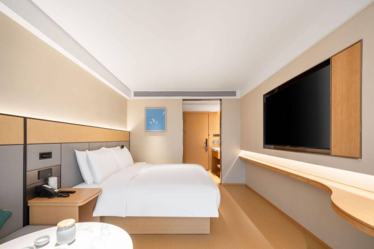 A modern hotel room with a large bed, a bedside table with a phone and a lamp, a flat-screen TV mounted on the wall, and a small round table with a cup and saucer.