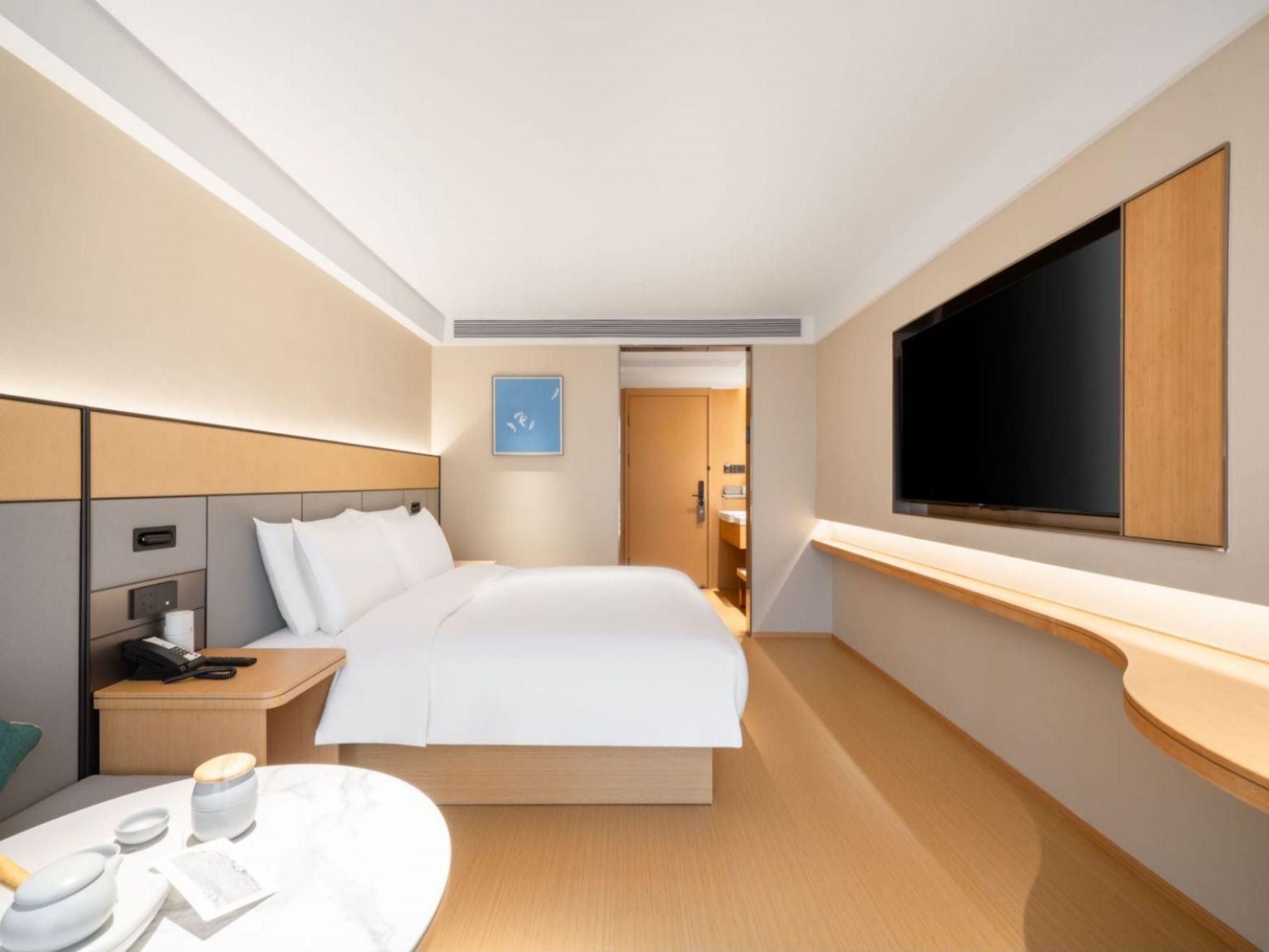 Una habitación de hotel moderna con una cama grande, una mesita de noche con teléfono y lámpara, un televisor de pantalla plana en la pared y una pequeña mesa redonda con una taza y plato.