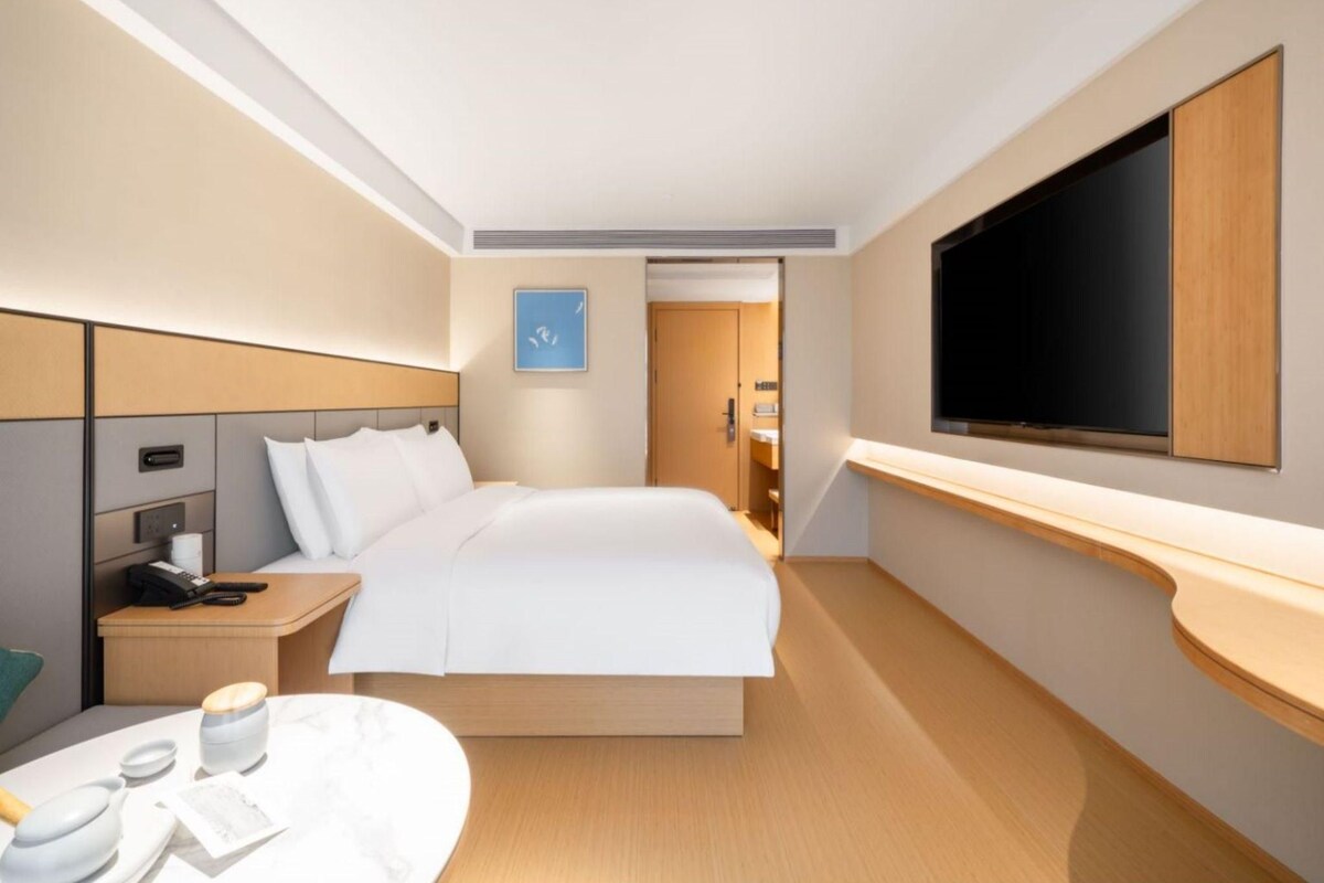 Een moderne hotelkamer met een groot bed, een nachtkastje met telefoon en lamp, een flatscreen televisie aan de wand en een klein rond tafeltje met kop en schotel.