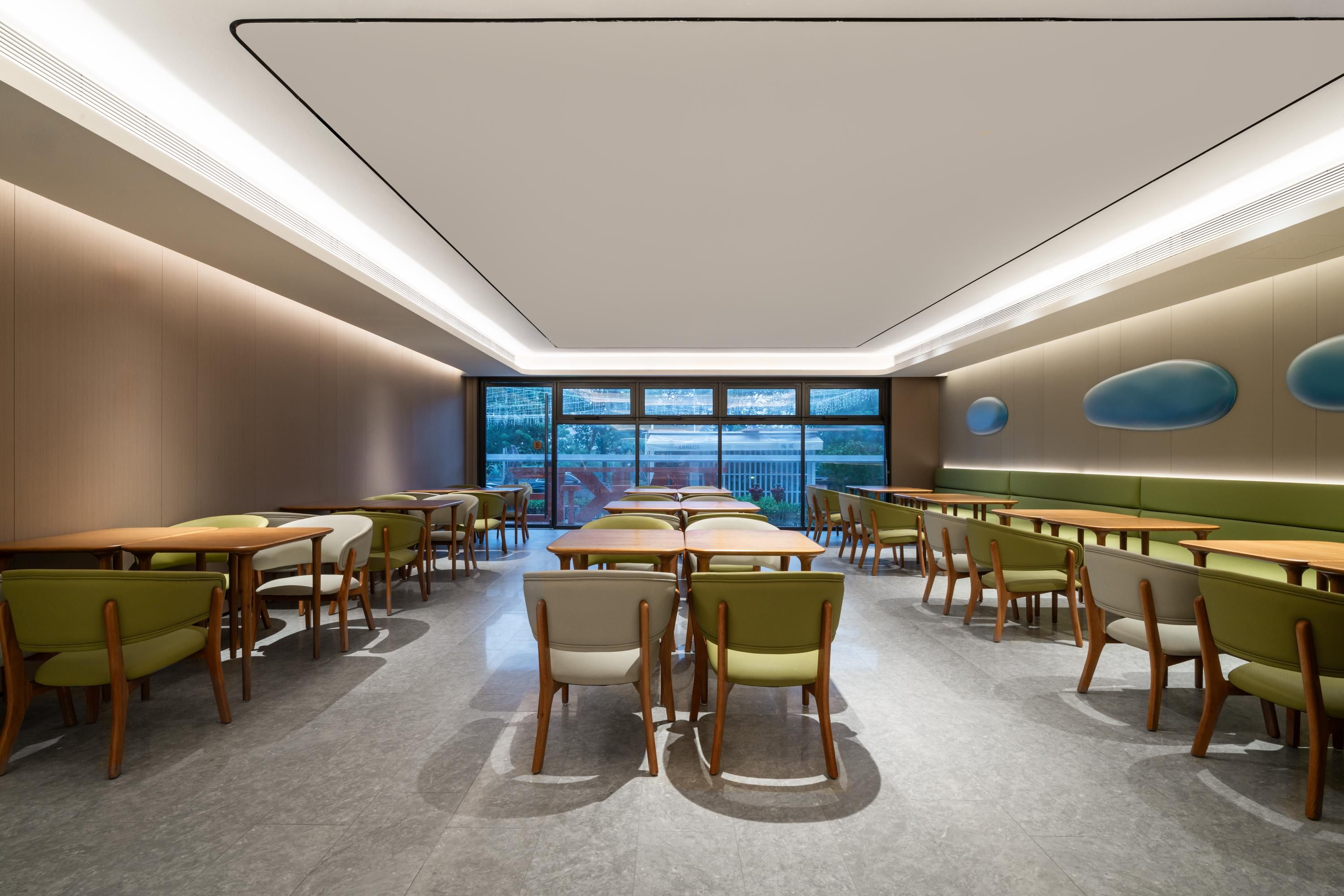 A modern dining area with tables and chairs arranged in rows, large windows providing a view of the outside, and a ceiling with recessed lighting.
