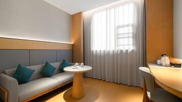 A modern hotel room with a grey sofa, teal cushions, a round wooden table, and a large window with sheer curtains.
