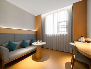 A modern hotel room with a grey sofa, teal cushions, a round wooden table, and a large window with sheer curtains. - Ji Guangzhou Liwan TCL Yunfeng Technology Park (Guangzhou)