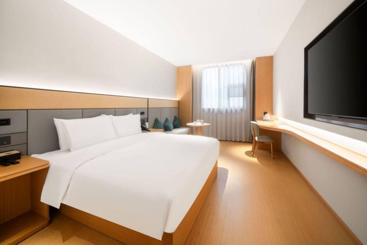 A modern hotel room with a large bed, a desk, a TV, and a window with curtains.