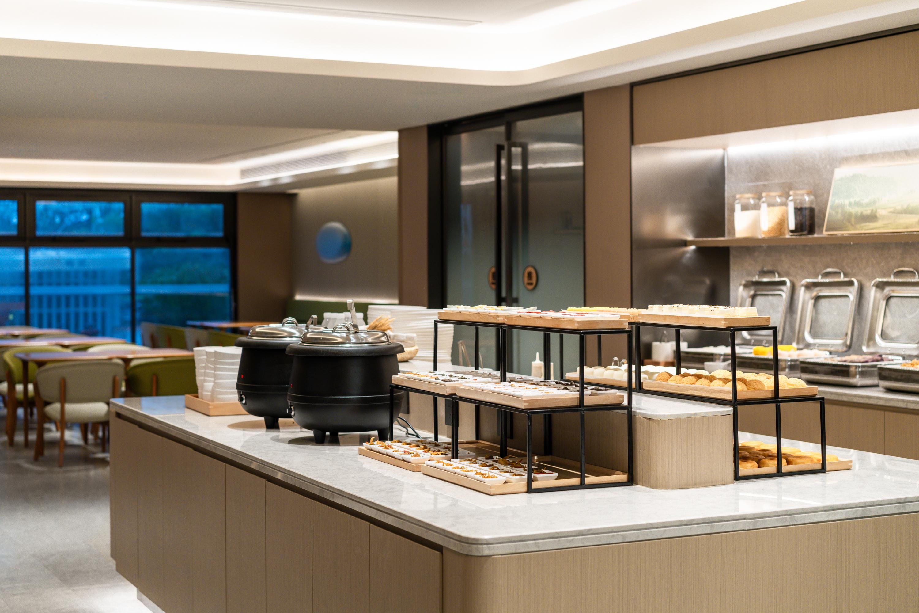 A modern breakfast buffet with multiple serving stations, a coffee machine, and a view of the outdoors through large windows.