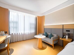 Ein modernes Hotelzimmer mit einem Holztisch, einem runden Tisch, einer Couch mit Kissen, einem Nachttisch mit Telefon und einem großen Fenster mit Vorhängen.