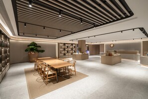 Een moderne hotellobby met een centrale houten tafel en stoelen, een grote potplant en een receptiebalie.