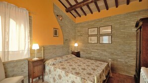 A bedroom with a bed, two bedside tables, a chair, a window with curtains, and a stone wall with framed pictures. - DEPENDANCE ON THE GARDEN (Uncinano, Spoleto)