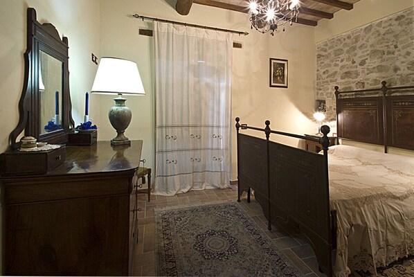 A bedroom with a wooden bed, a nightstand, a dresser, a mirror, a lamp, a window with a white curtain, and a chandelier.