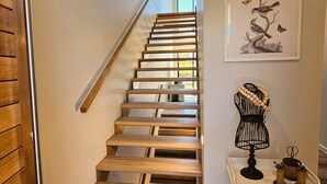 Puinen portaikko, jossa on metallinen kaide, seinällä oleva kehystetty lintukuvitus sekä pieni pöytä, jonka päällä on musta metallirunkoinen lamppu ja koriste-esineitä.