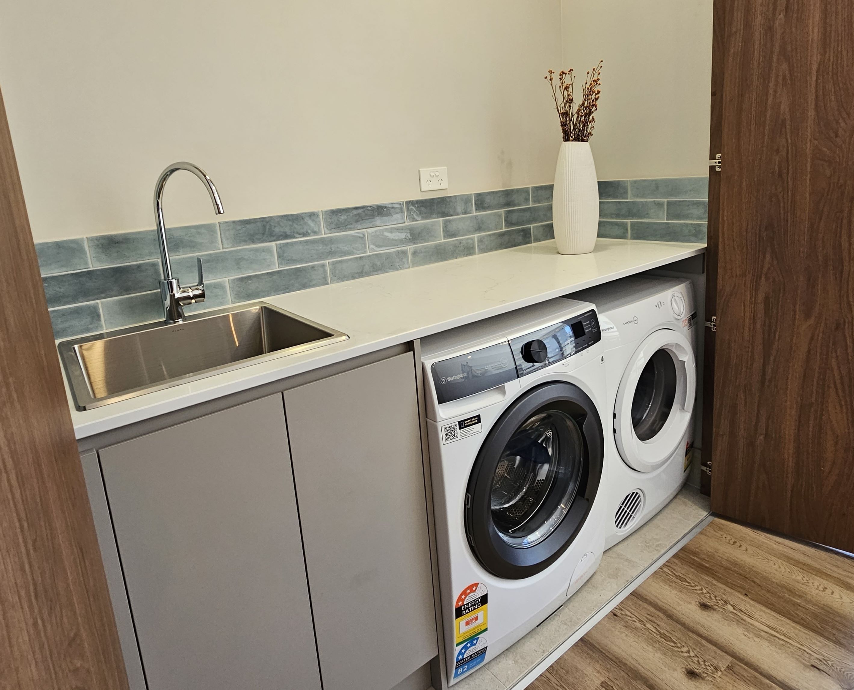 A compact laundry room with a stainless steel sink, a washing machine, and a vase with dried flowers on the countertop.