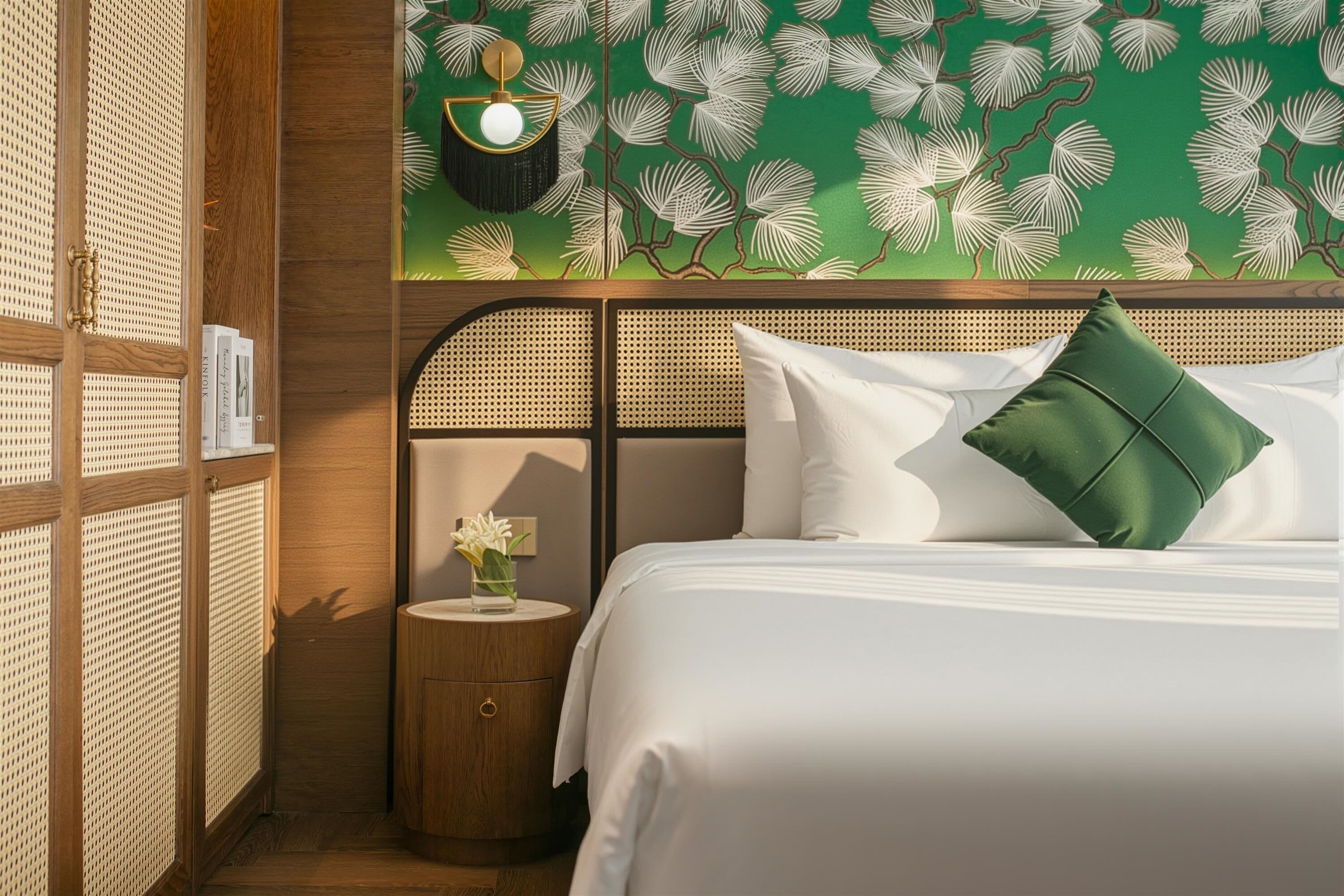 A neatly made bed with white and green pillows, a round wooden nightstand with a vase of flowers, and a wall with a leafy pattern.