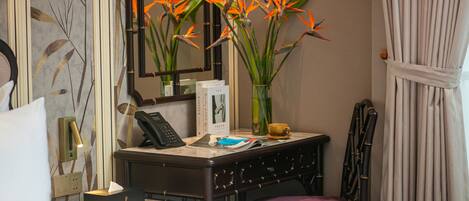Habitación de hotel con una cama, un escritorio con teléfono, un florero con flores, un espejo y una silla.