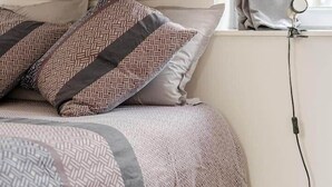 A bedroom with a bed, patterned pillows, a window with curtains, and a visible electrical outlet. - Welcome to Marie & Guillaume's 2* rated home (Carentan-les-Marais)