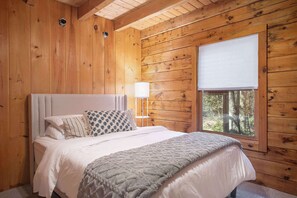 Un dormitorio de cabaña de madera con cama, ventana con vista a árboles y mesita de noche con lámpara.