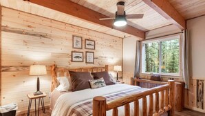 A cozy wooden cabin bedroom with a bed, two bedside lamps, a window with a view of trees, and a ceiling fan. - Barratt Lodge | Hot Tub | 35 mins to Yellowstone (Island Park)