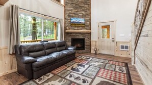 A cozy living room with a stone fireplace, a leather sofa, and a large window overlooking a forest. - Barratt Lodge | Hot Tub | 35 mins to Yellowstone (Island Park)