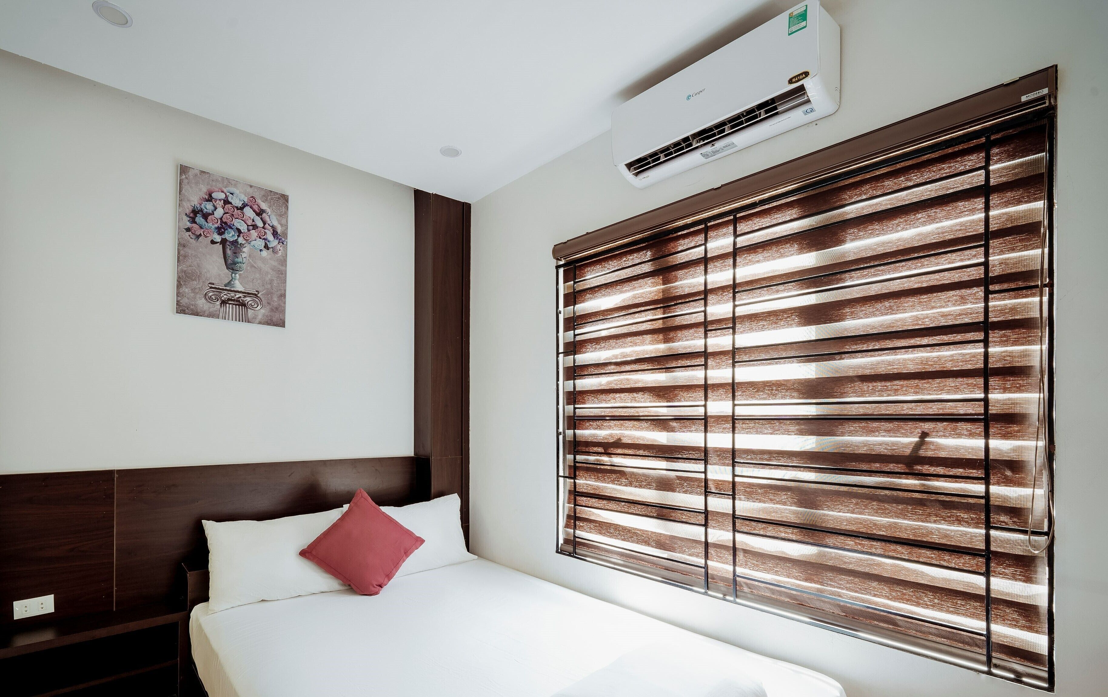 A hotel room with a bed, a nightstand, a red pillow, a wall-mounted air conditioner, and a window with horizontal blinds.