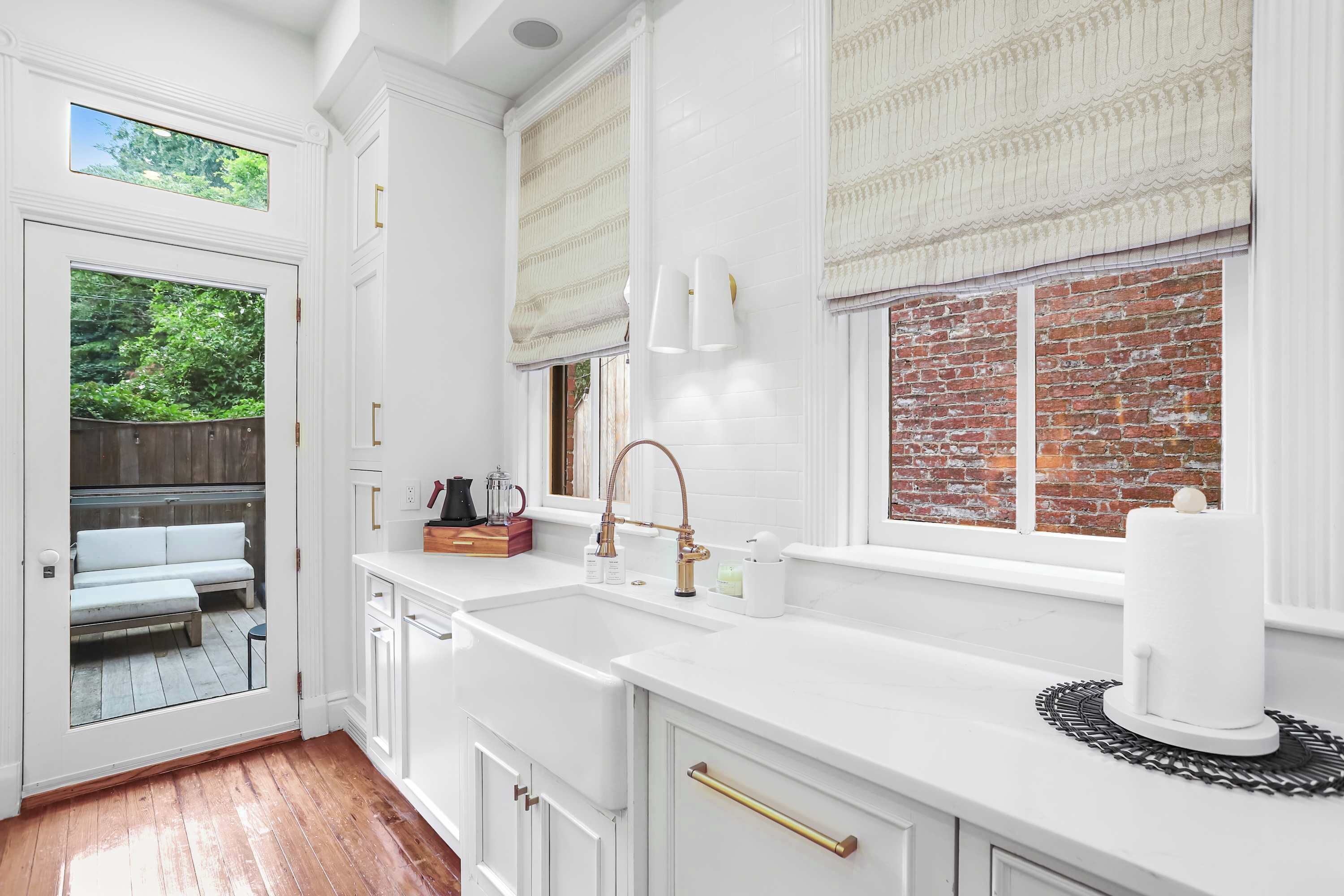 A kitchen with white cabinetry, a large window with a view of a brick wall and greenery, and a built-in sink with a gold faucet.