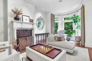A living room with a fireplace, a chessboard, a sofa, and large windows with a view of greenery.