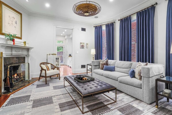 A living room with a fireplace, a sofa, a coffee table, and a view of a brick wall through blue curtains.
