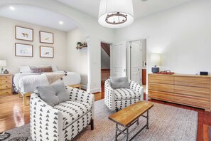A bedroom with a bed, two patterned armchairs, a wooden bench, a dresser, and a hallway leading to a staircase.